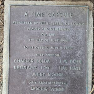 A weathered bronze plaque set in a concrete border on the ground. The text reads: "A TIME CAPSULE ASSEMBLED BY THE CITIZENS OF LOMITA, SEALED AND DEDICATED ON THE 5TH DAY OF JULY 1976, TO BE OPENED JULY 4, 2026." It lists City Council members Charles Belba, Jim Cole, Leonard Loy, Hal Hall, Jerry Moore, and City Administrator Morris Vance.