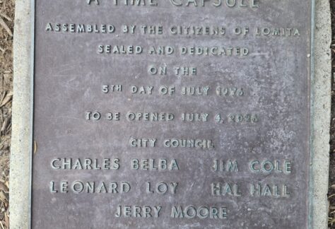 A weathered bronze plaque set in a concrete border on the ground. The text reads: "A TIME CAPSULE ASSEMBLED BY THE CITIZENS OF LOMITA, SEALED AND DEDICATED ON THE 5TH DAY OF JULY 1976, TO BE OPENED JULY 4, 2026." It lists City Council members Charles Belba, Jim Cole, Leonard Loy, Hal Hall, Jerry Moore, and City Administrator Morris Vance.