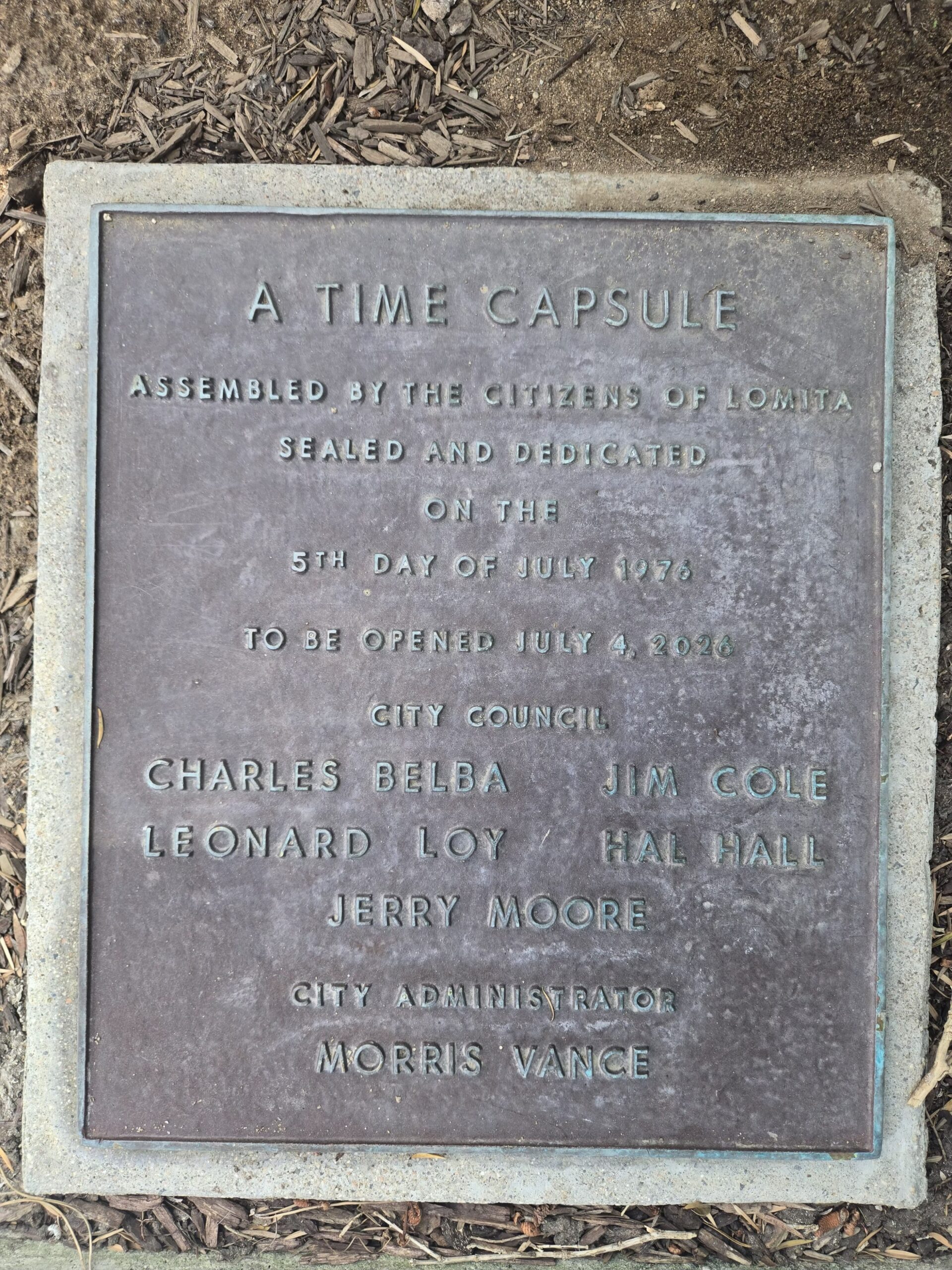 A weathered bronze plaque set in a concrete border on the ground. The text reads: "A TIME CAPSULE ASSEMBLED BY THE CITIZENS OF LOMITA, SEALED AND DEDICATED ON THE 5TH DAY OF JULY 1976, TO BE OPENED JULY 4, 2026." It lists City Council members Charles Belba, Jim Cole, Leonard Loy, Hal Hall, Jerry Moore, and City Administrator Morris Vance.