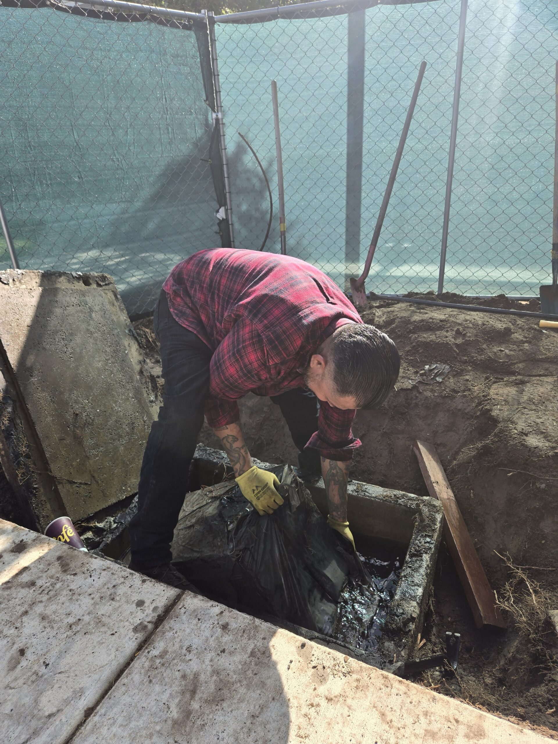 A man in a red plaid shirt and work gloves bends over an open underground vault, lifting a heavy object wrapped in a wet, muddy black plastic bag out of the standing water inside.