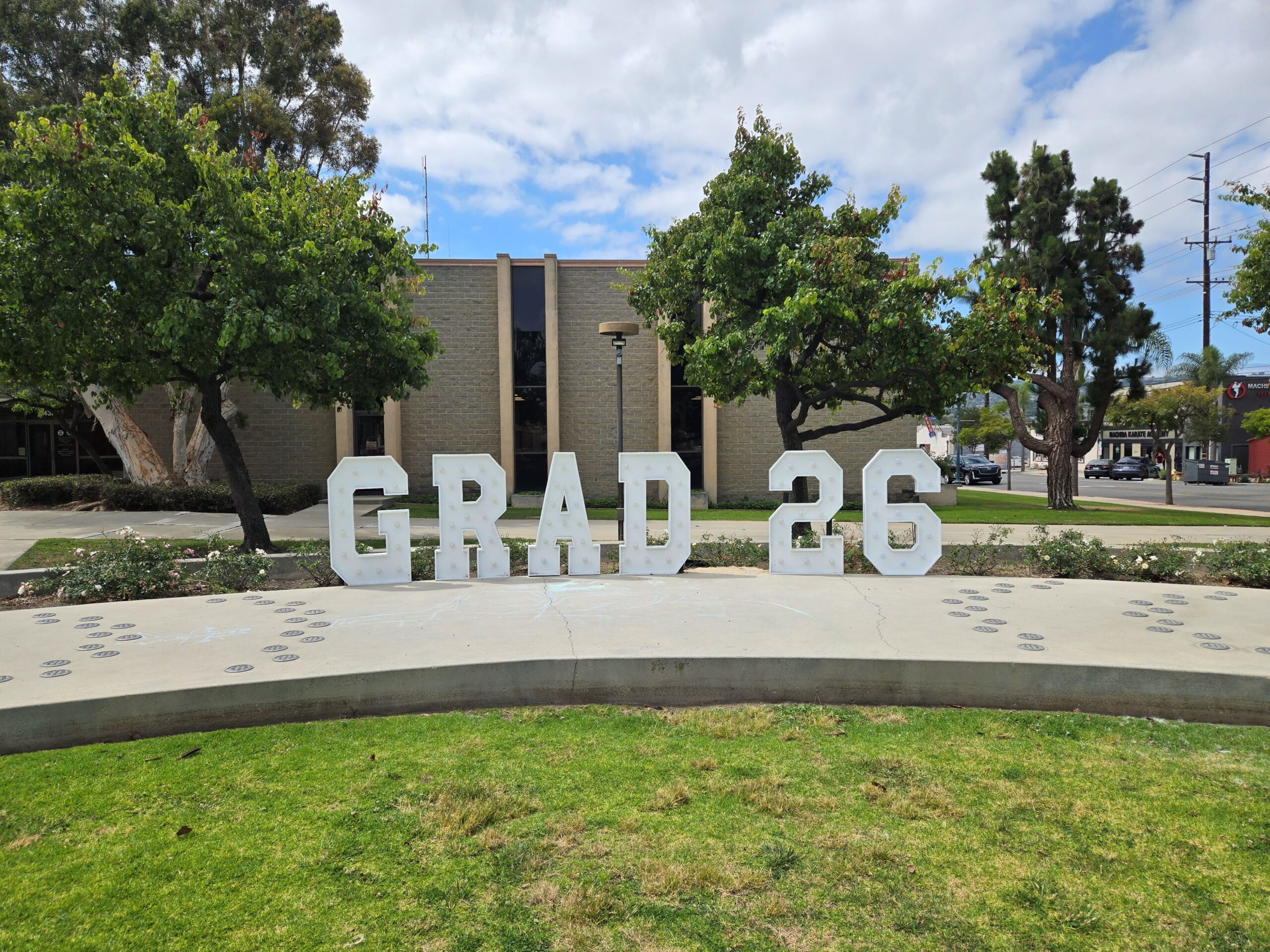 Grad Sign 2 Large, white marquee letters spelling out 'GRAD 26' with built-in lightbulbs, set up on a concrete walkway on Lomita's City Hall campus. In the background are green trees and a modern brick building under a partly cloudy blue sky.