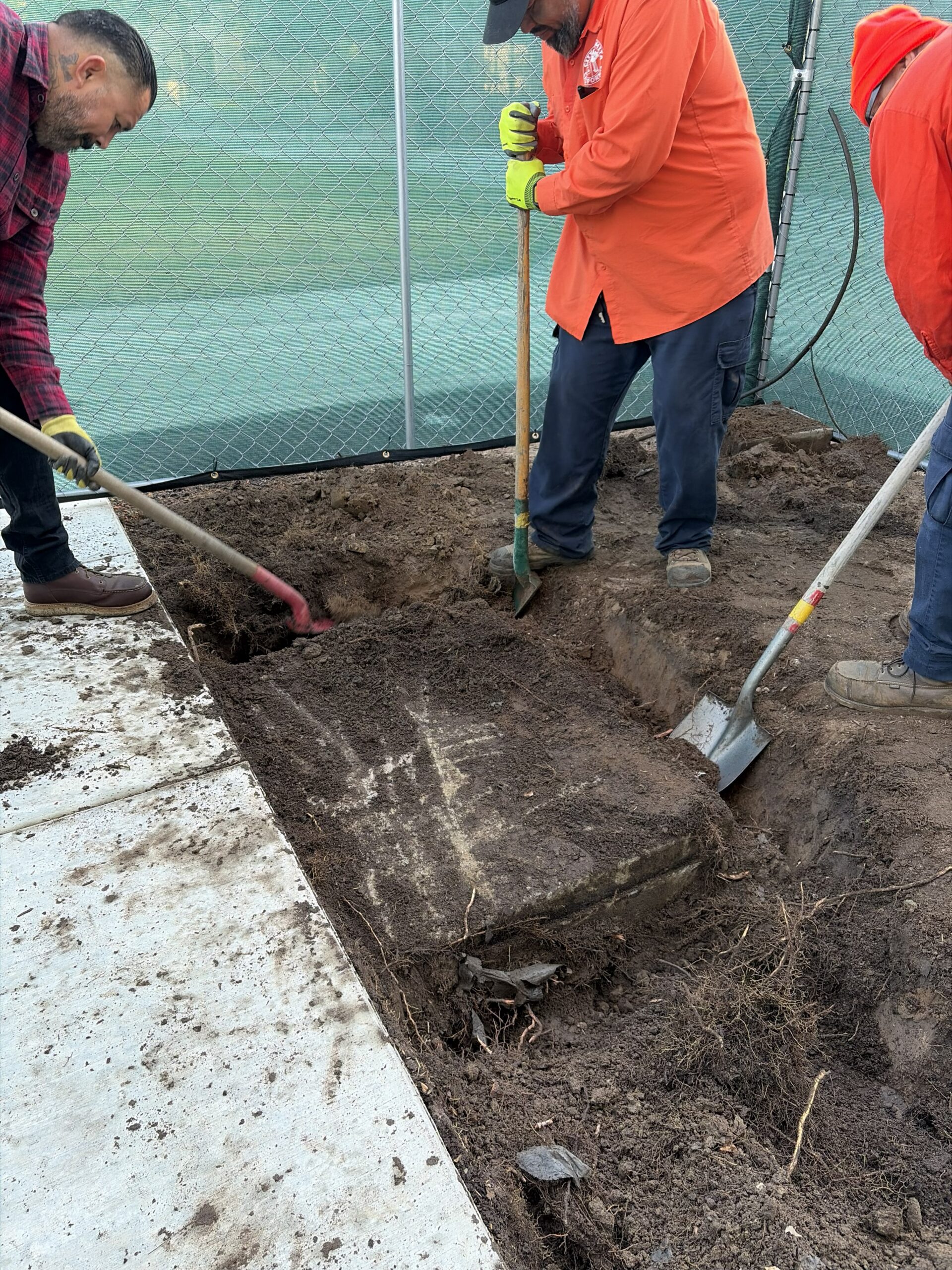 Three city workers using shovels to dig up dirt around a large concrete slab embedded in the ground next to a newly poured concrete sidewalk, surrounded by construction fencing.