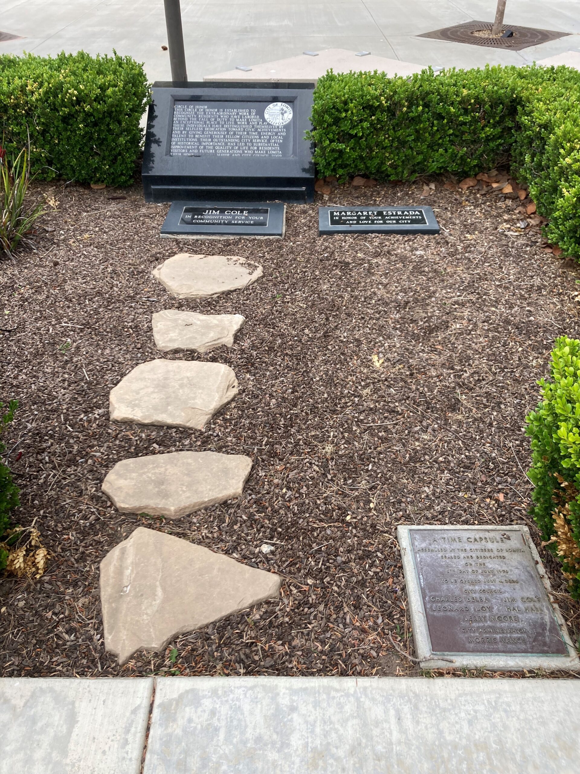 A stone-lined garden bed in Lomita, the Circle of Honor, featuring black granite plaques dedicating the monument to residents Jim Cole and Margaret Estrada. Five stepping stones lead through the mulch toward the plaques, and a bronze time capsule plaque from 1976 sits in the bottom right corner.