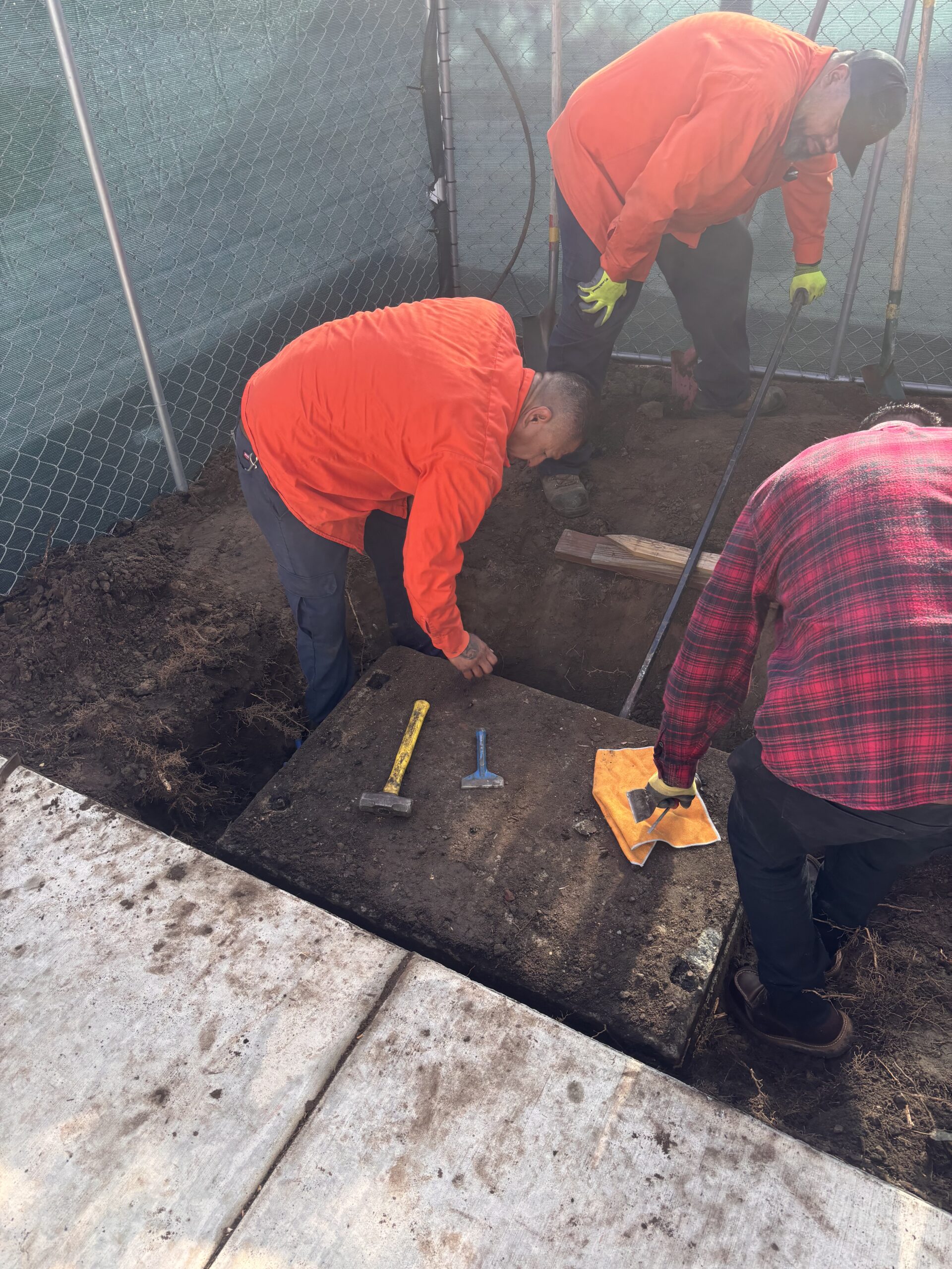 Overhead view of three workers clearing dirt off the top of a heavy, rectangular concrete slab lid using hand tools, a hammer, and a towel.