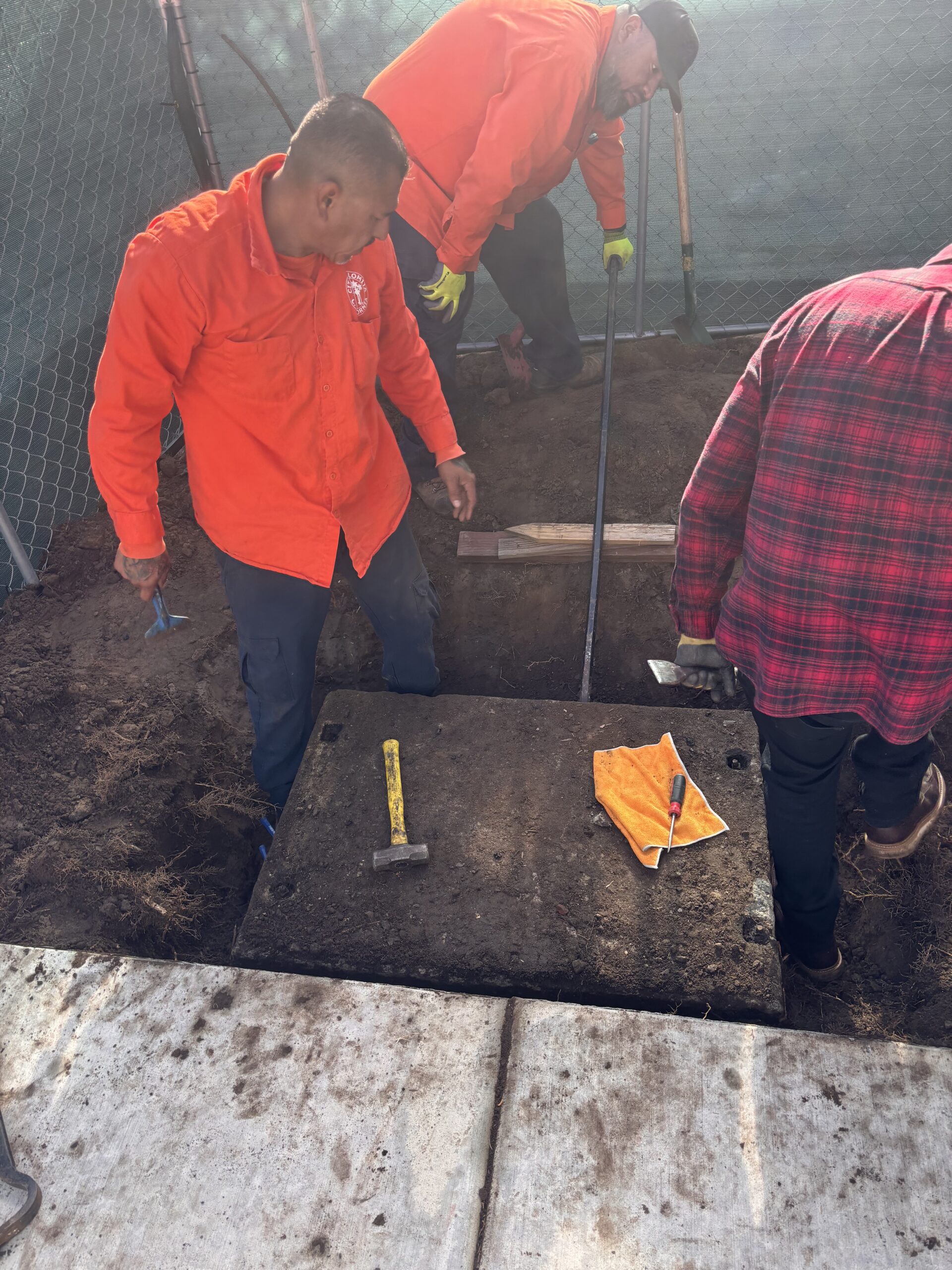 Workers using a long metal pry bar and wooden blocks to leverage and loosen the heavy concrete lid of a buried vault.