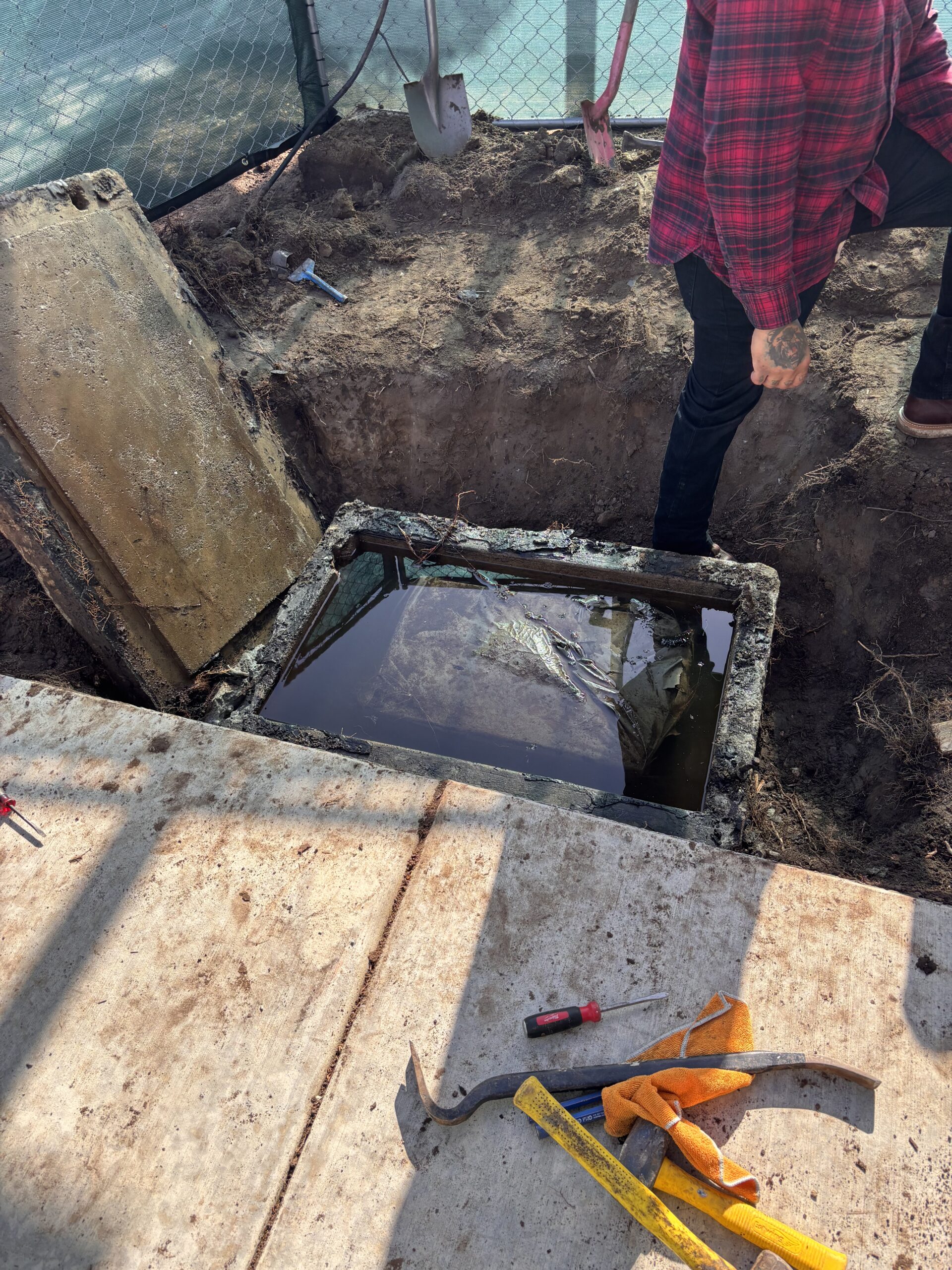 A wide view of the uncovered concrete vault with its lid propped open, showing the water-logged interior and tools scattered on the adjacent sidewalk.