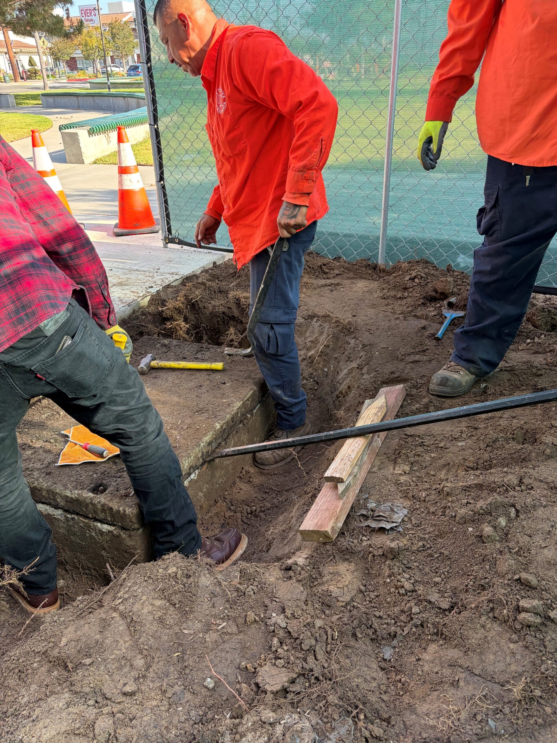 Workers using a crowbar, pry bar, and hand tools to carefully dig out a trench around the edge of a large, flat concrete lid buried in the dirt.