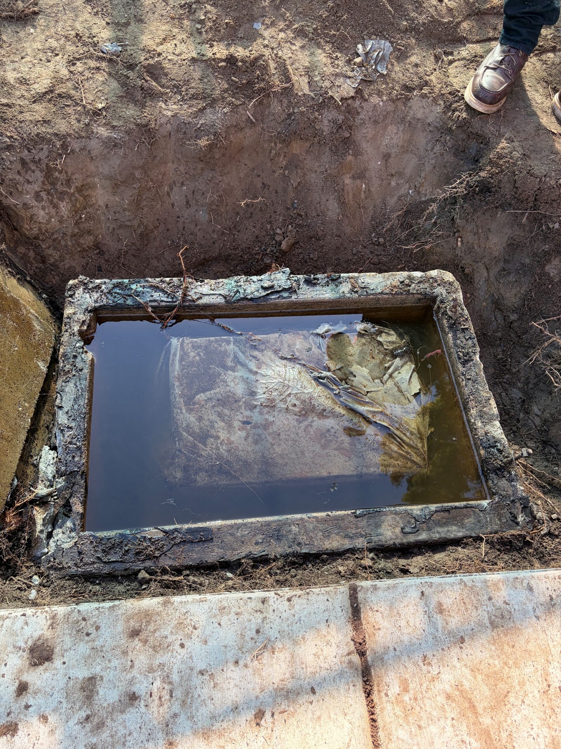 A top-down view of an opened underground concrete vault filled with dark water, revealing a large object wrapped in protective plastic submerged inside.
